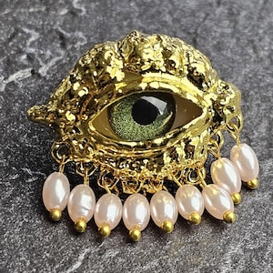 Könnte beinhalten: Eine goldfarbene Brosche in Form eines Auges mit strukturierter, metallischer Oberfläche. Die Mitte zeigt eine grüne Iris und eine schwarze Pupille. Am unteren Rand hängen neun kleine, ovale, schimmernde weiße Perlen an goldenen Ringen.