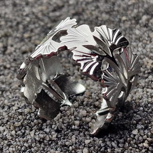 Puede incluir: Un anillo plateado con un diseño detallado de hojas. El anillo abierto presenta múltiples hojas estilizadas, creando una estética inspirada en la naturaleza. El anillo está sobre un fondo gris texturizado.
