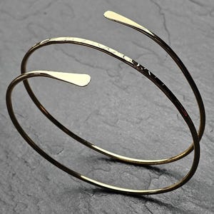 May include: A gold-coloured arm cuff bracelet with a minimalist design. The open cuff features two curved bands that wrap around the arm, with flattened, paddle-shaped ends. The bracelet is displayed on a dark grey, textured surface.