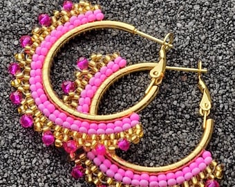 Pode incluir: Um par de brincos de argola com contas rosa e douradas. Os brincos são feitos de metal e têm a forma de argola. As contas são dispostas em um padrão que cria um efeito decorativo.