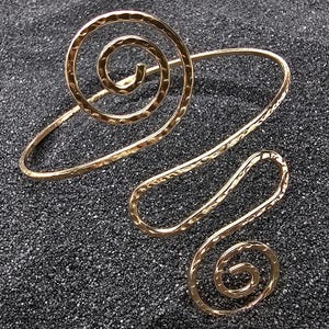 May include: A gold arm cuff with a hammered finish. The cuff has a large spiral design and a smaller spiral at the bottom, linked by a curved band. The jewellery is presented on a dark, textured surface.