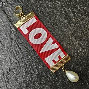 May include: A red ribbon pendant with the word "LOVE" in white block letters. The ribbon is trimmed with gold and has a gold loop at the top and a pearl drop at the bottom. The pendant is set against a dark, textured background.