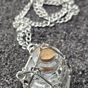 Peut inclure: Pendentif en forme de flacon en verre transparent avec bouchon en liège, enfermé dans une cage en métal argenté et suspendu à une chaîne assortie. Le collier est présenté sur une surface grise texturée.