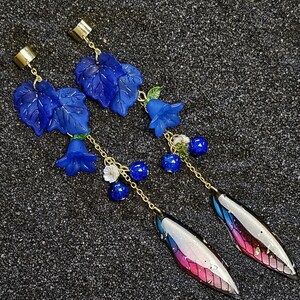 Può includere: Un paio di orecchini pendenti con dettagli di foglie e fiori blu. Gli orecchini hanno catene e ganci per le orecchie color oro. La parte inferiore degli orecchini presenta un design argento, rosa e blu.