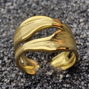 Op de afbeelding: Een gouden ring met een uniek, golvend ontwerp. De ring is gemaakt van twee met elkaar verweven metalen delen.