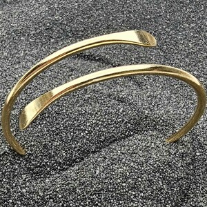 Könnte beinhalten: Ein goldfarbenes Armreif-Armband mit geschwungenem, offenem Design. Das Armband hat eine glatte, polierte Oberfläche und ist vor einem strukturierten, dunkelgrauen Hintergrund platziert. Die Enden des Armbands sind leicht abgeflacht.
