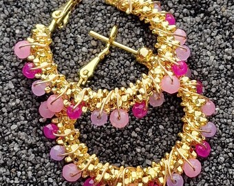 Pode incluir: Brincos de argola dourados com contas rosa e roxas. Os brincos são feitos de fio e têm um visual delicado e feminino.