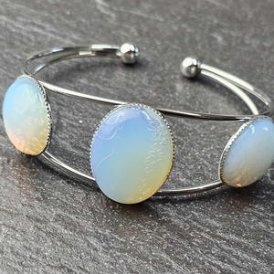 Pode incluir: Pulseira de punho prateada com três pedras ovais de opala iridescentes. As pedras têm uma cor branca perolada e amarelo claro. A pulseira é aberta com pequenas bolas prateadas nas extremidades.