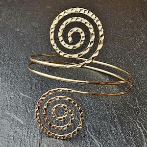 Peut inclure: Manchette de bras dorée avec un motif en spirale. Le bracelet est composé de deux bandes qui s'enroulent autour du bras, avec un détail en spirale texturé en haut et un accent en spirale séparé. Le bijou a une finition martelée.