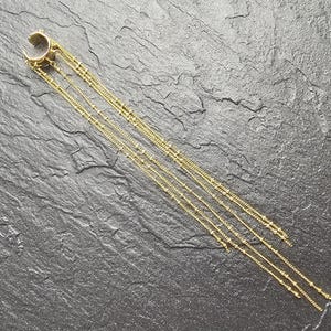 May include: A gold-toned ear cuff featuring a circular band with an open gap, connected to four long, delicate dangling chains. Each chain is adorned with small, evenly spaced metallic beads, creating a layered, cascading effect in length.