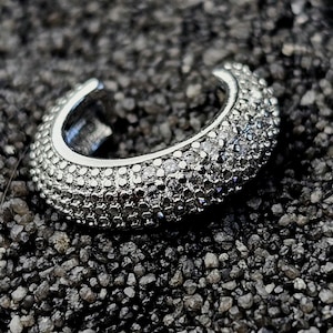 Può includere: Un orecchino a cerchio d'argento, incrostato con numerose piccole pietre scintillanti. Il gioiello è curvo e progettato per avvolgere l'orecchio. Il cerchio è posto su uno sfondo scuro e strutturato, creando un contrasto che ne esalta la brillantezza.