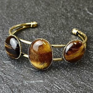 Könnte beinhalten: Ein goldfarbenes Armreif-Armband mit drei ovalen Tigeraugensteinen. Die Steine sind braun und goldfarben, und das Armband liegt auf einer dunkelgrauen, strukturierten Oberfläche. Das Armband ist ein Schmuckstück.