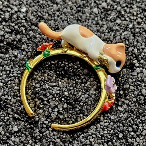 May include: A gold-tone open ring featuring a small, white and orange cat figurine resting on the band. The ring is decorated with small, colourful enamel flowers and green leaves along the sides, creating a detailed and vibrant accessory piece.