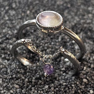 Może przedstawiać: Dwa srebrne pierścionki. Górny pierścionek ma okrągły, jasny kamień. Dolny pierścionek jest połączony z górnym delikatnym łańcuszkiem i ma mały, fioletowy kamień w kształcie serca. Pierścionki są na ciemnym, teksturowanym tle.