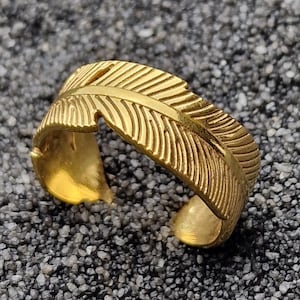 Op de afbeelding: Een goudkleurige veering. De ring is aan de onderkant open en wikkelt zich om de vinger. Het veermotief heeft gedetailleerde lijnen, waardoor het er realistisch uitziet. De ring staat tegen een donkere, getextureerde achtergrond.