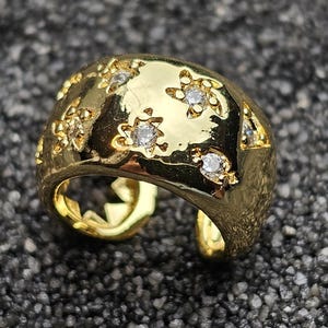Könnte beinhalten: Ein goldfarbener Ring mit einem einzigartigen Design. Der Ring hat sternförmige Aussparungen und ist mit kleinen, klaren Edelsteinen besetzt. Der Ring liegt auf einer strukturierten, dunkelgrauen Oberfläche.