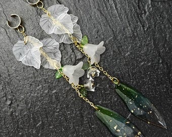 May include: A pair of long, dangling earrings featuring translucent white leaf and flower accents, connected by gold-colored chains. The bottom of each earring has a dark green, teardrop-shaped element with gold flecks. The earrings are set against a dark, textured background.