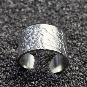 Può includere: Un anello largo e aperto in argento con una superficie strutturata. L'anello presenta un motivo organico, simile alla corteccia, ed è posto su uno sfondo scuro e maculato. Il design dell'anello è moderno e minimalista.