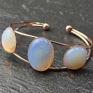Pode incluir: Uma pulseira de punho em ouro rosa com três pedras ovais opalescentes. As pedras exibem cores iridescentes de azul, amarelo e branco. A pulseira tem um design aberto, com as pedras emolduradas em uma estrutura delicada.