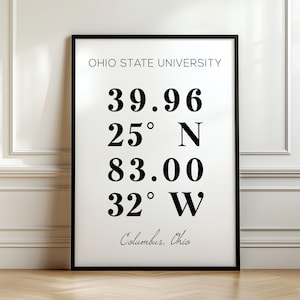 May include: A framed print with the text "OHIO STATE UNIVERSITY" at the top. Below are coordinates and the city name "Columbus, Ohio". The print is in a black frame and hangs on a white wall.