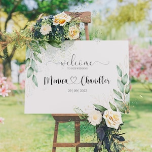 May include: A white wedding welcome sign with green and gold floral accents. The sign reads "Welcome to our wedding, Monica & Chandler, 29 | 09 | 2022".