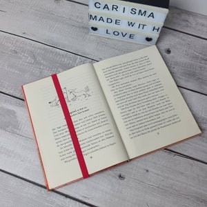 May include: A close-up of an open book with a red bookmark. The book is titled "Carisma Made With Love" and features a black and white illustration of two dogs. The text on the page is in German.