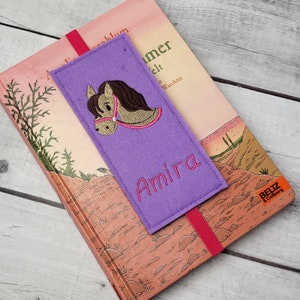 May include: Purple felt bookmark with a brown horse head embroidered on it. The bookmark has the name "Amira" embroidered in pink lettering.