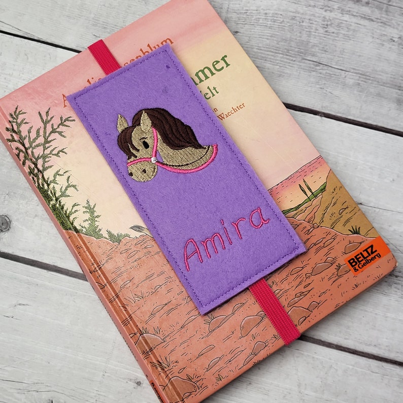 May include: Purple felt bookmark with a brown horse head embroidered on it. The bookmark has the name "Amira" embroidered in pink lettering.