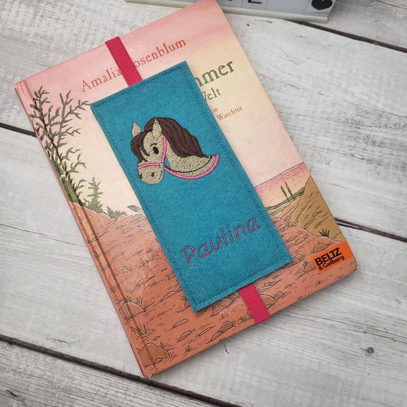 May include: A teal felt bookmark with a pink elastic band. The bookmark features an embroidered brown horse with a pink bridle and the name "Paulina" embroidered in pink.