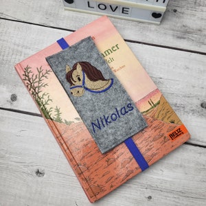 May include: A gray felt bookmark with a blue elastic band. The bookmark has an embroidered brown horse with a blue bridle and the name "Nikolas" in blue thread.