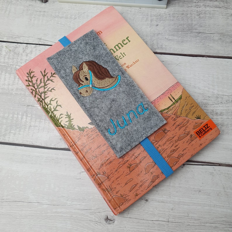May include: A gray felt bookmark with a blue ribbon. The bookmark has an embroidered brown horse with a blue bridle and the name "Juna" in blue thread.