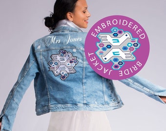 Pode incluir: Uma jaqueta de ganga azul claro com um design bordado branco nas costas. O design apresenta dois pássaros e o texto "Bride Jacket Embroidered". A jaqueta tem uma etiqueta branca com o texto "Mrs. Jones" nas costas. A modelo está usando uma saia branca.
