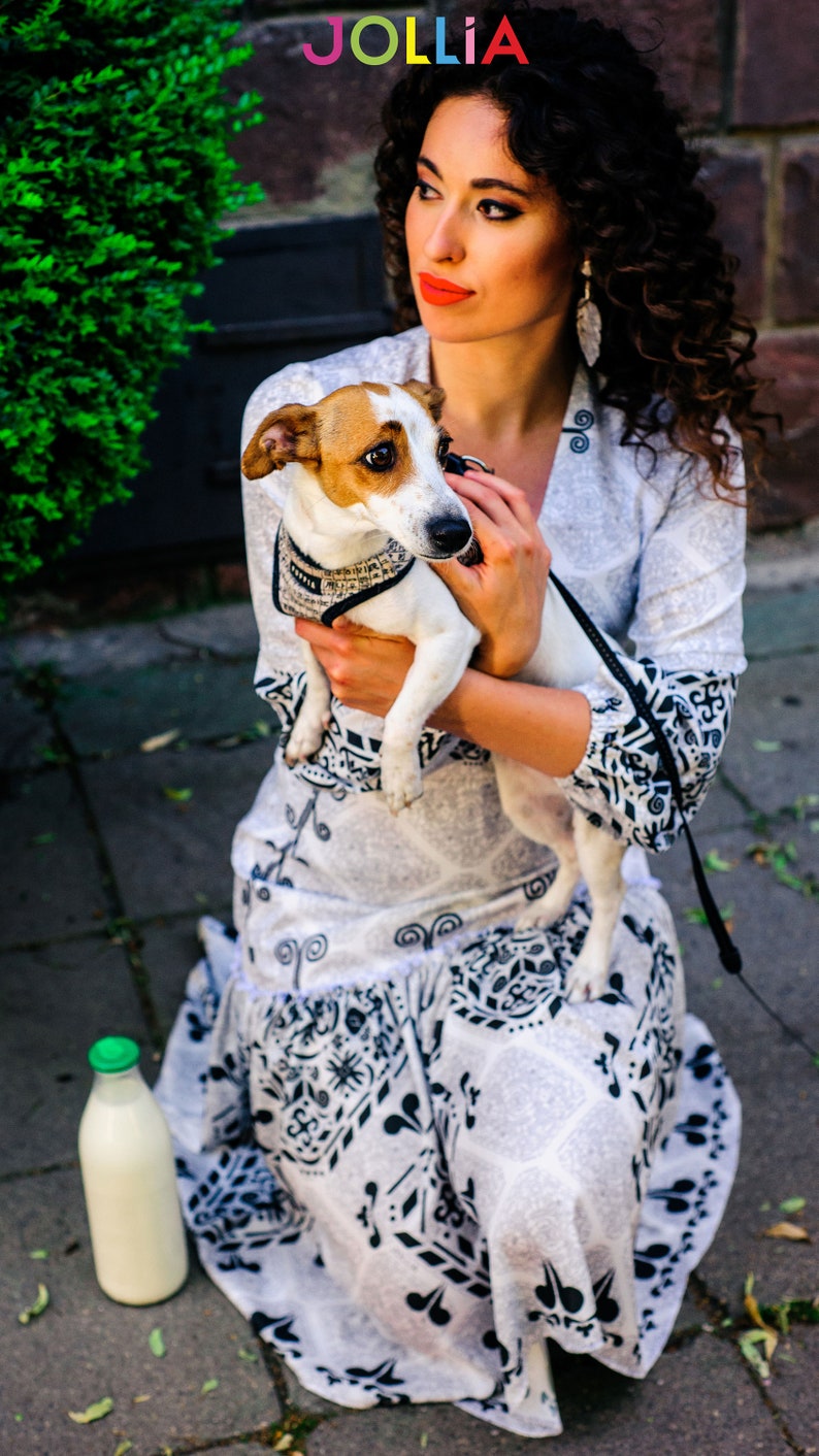 Op de afbeelding: Een vrouw met krullend bruin haar houdt een kleine bruin-witte hond vast. Ze draagt een witte jurk met een zwart-wit patroon. De hond draagt een zwart harnas. De vrouw zit op een stoep met een fles melk naast haar.