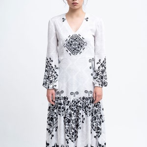 Op de afbeelding: Een witte midi-jurk met zwarte geometrische patronen. De jurk heeft lange mouwen en een V-hals. Het model draagt zwarte enkellaarsjes met witte tenen.