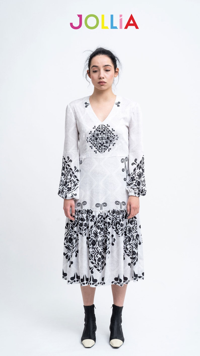 Op de afbeelding: Een witte midi-jurk met zwarte geometrische patronen. De jurk heeft lange mouwen en een V-hals. Het model draagt zwarte enkellaarsjes met witte tenen.