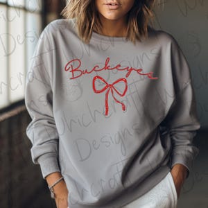 Könnte beinhalten: Ein graues Sweatshirt mit dem Wort "Buckeyes" in roter Schrift und einer roten Schleifen-Grafik. Das Sweatshirt hat einen Rundhalsausschnitt und lange Ärmel. Das Design befindet sich auf der Vorderseite.