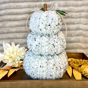 May include: A stacked pumpkin decoration made of cream-colored speckled yarn with a ribbed knit texture. The top features a brown cinnamon stick stem, a white sheer ribbon bow, a piece of twine, and a small green leaf accent.