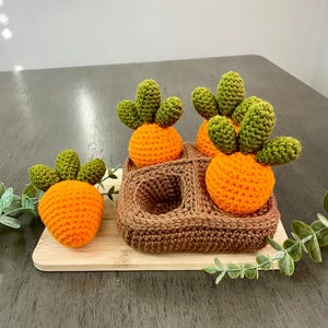 May include: Crocheted orange carrots with green tops, displayed on a wooden tray and a brown crocheted basket. The carrots are handmade and have a textured appearance. The arrangement is set against a neutral background, suggesting a rustic or handmade theme.