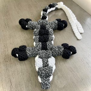 May include: A handmade crochet alligator toy in black, white, and gray. The alligator has a textured body, four legs, and a long tail. The toy is lying on a light-colored surface.