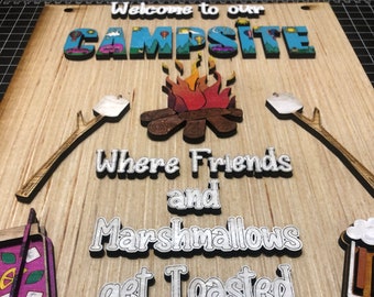 May include: Wooden sign with the words "Welcome to our Campsite" and "Where Friends and Marshmallows get Toasted." Features a campfire, marshmallows, and an arrow. The sign is made of wood.