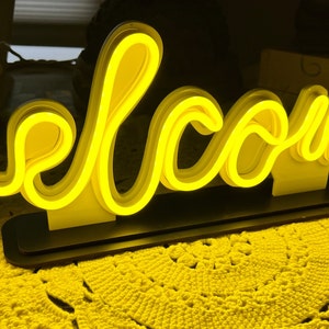 May include: A neon sign displaying the word "welcome" in a flowing, cursive font. The sign emits a bright yellow glow and is mounted on a black base. The background features a yellow crocheted surface.