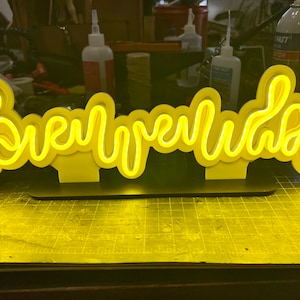 May include: A yellow neon sign that reads "Brewery Labs" in a cursive font. The sign is layered and sits on a black base, casting a yellow glow. The background is a cluttered workspace.