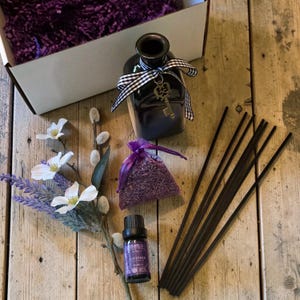 Peut inclure: Un flacon diffuseur en verre noir avec un ruban à carreaux noir et blanc et une breloque clé en métal, un petit sachet en organza violet rempli de lavande séchée, une fiole d'huile de lavande et des bâtonnets diffuseurs noirs près d'une tige fleurie.