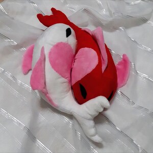 Könnte beinhalten: Zwei Plüschfischspielzeuge, eines weiß mit rosa Akzenten und das andere rot mit rosa Akzenten, kuscheln sich auf einem weißen Hintergrund aneinander.