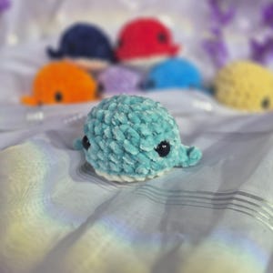 May include: A collection of small, plush, crocheted whale toys in various colours including light blue, orange, dark blue, red, purple, and yellow. Each whale features a soft, textured yarn finish, black plastic eyes, and a white underbelly.