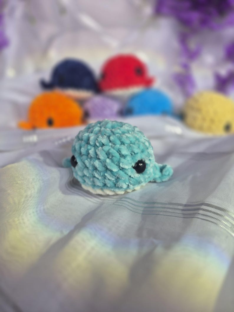 May include: A collection of small, plush, crocheted whale toys in various colours including light blue, orange, dark blue, red, purple, and yellow. Each whale features a soft, textured yarn finish, black plastic eyes, and a white underbelly.
