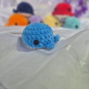 May include: A small, blue, crocheted whale plush toy with a soft, textured yarn finish and a single black plastic eye. In the background, several other similar whale plush toys in various colours, including red, orange, yellow, and teal, are visible.