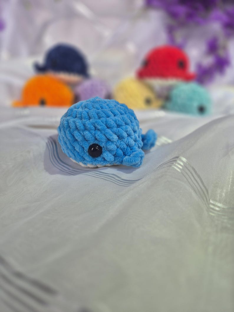 May include: A small, blue, crocheted whale plush toy with a soft, textured yarn finish and a single black plastic eye. In the background, several other similar whale plush toys in various colours, including red, orange, yellow, and teal, are visible.