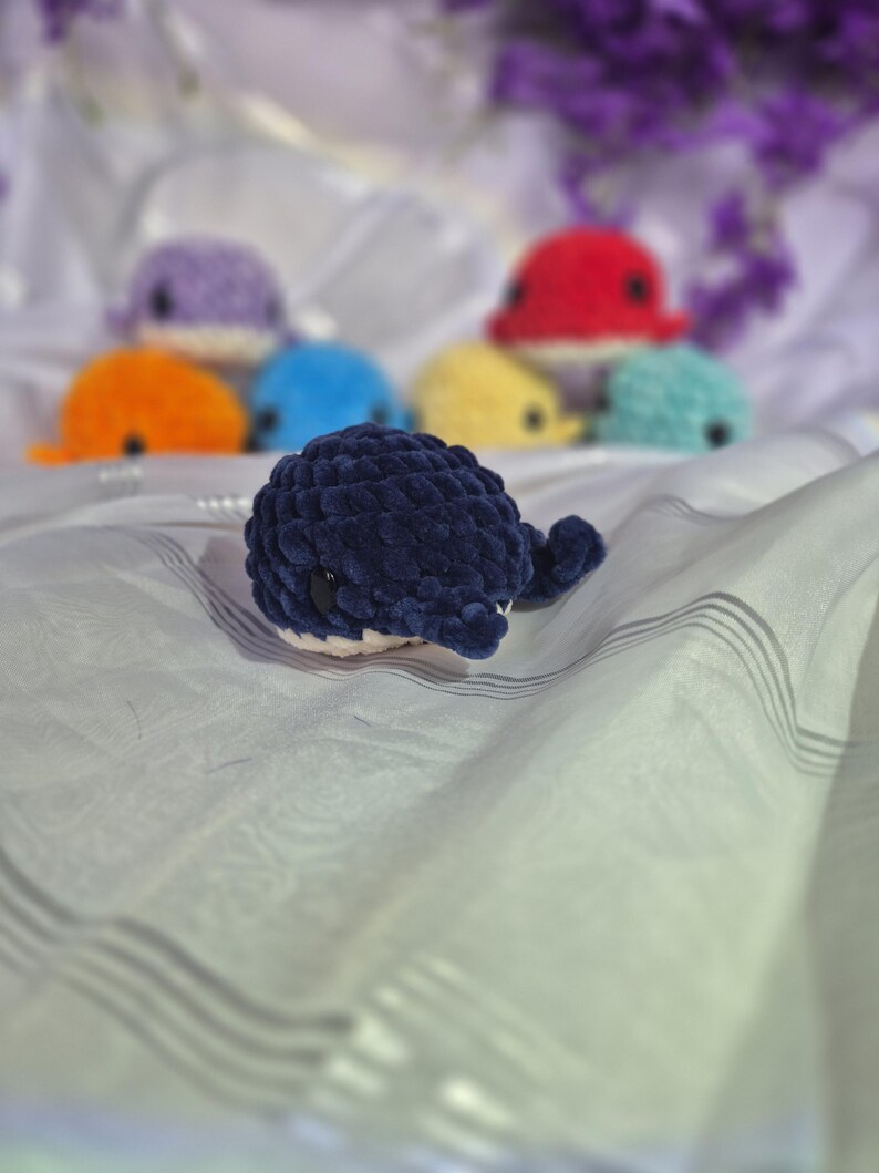 May include: A dark blue crocheted whale plush toy with a white underbelly and black plastic eyes. In the background, several other small crocheted whale plush toys in various colours including purple, orange, red, yellow, and light blue are visible.