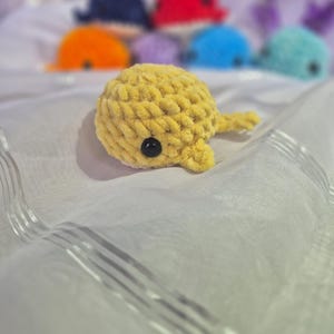 May include: A small, yellow, crocheted whale plushie made from soft, chunky yarn. The whale features a rounded body, a small tail, and a single visible black plastic eye on its side. Several other colourful crocheted whale plushies are blurred in the background.